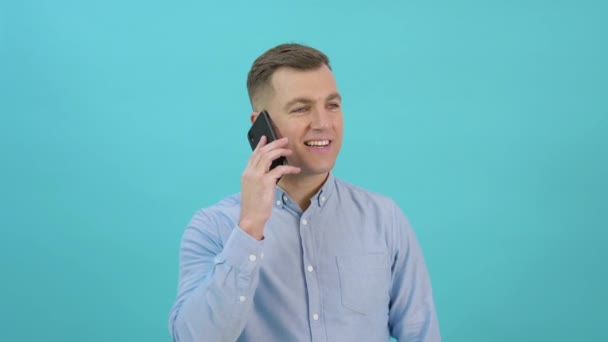 Caucásico hombre de mediana edad en una camisa azul levanta un teléfono inteligente a su oído y habla. Trabajador de oficina teniendo una conversación telefónica frente a un fondo azul brillante — Vídeos de Stock