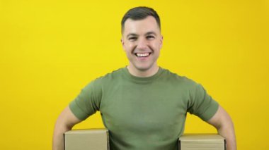 Caucasian middle-aged man in a green T-shirt holding two craft cardboard boxes. The guy is standing in front of a yellow background with 2 parcels in his hands. Yellow-green color scheme