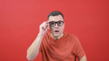 A man in glasses looks into the camera as if he cant see well. A guy in red clothes stands in front of a red background and expresses surprise