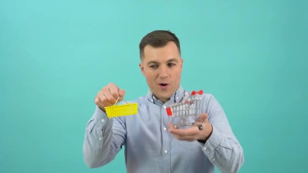 A customer man in a blue shirt happily demonstrates a toy basket and shopping cart in his hands. Business man on a bright blue background having fun with the attributes of shopping in the supermarket — Stok video