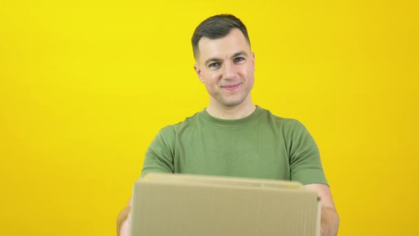 Young delivery man courier in a green T-shirt lifts a craft cardboard box in front of him. The guy is standing in front of a yellow background with a parcel in his hands — 图库视频影像