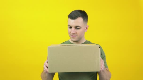 Caucasian middle-aged man in a green T-shirt lifts a craft cardboard box in front of him. The guy is standing in front of a yellow background with a parcel in his hands — стокове відео