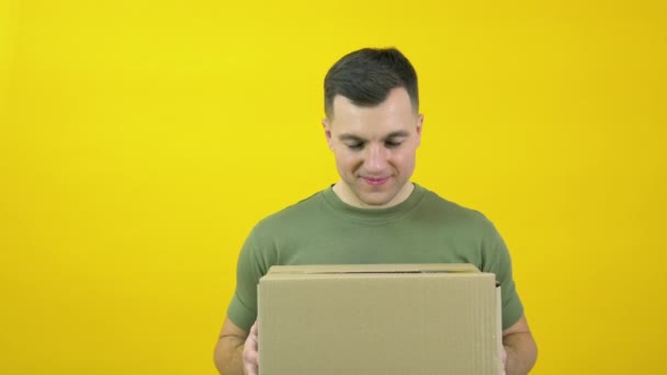 Young delivery man courier in a green T-shirt lifts a craft cardboard box in front of him. The guy is standing in front of a yellow background with a parcel in his hands. Yellow-green color scheme — ストック動画