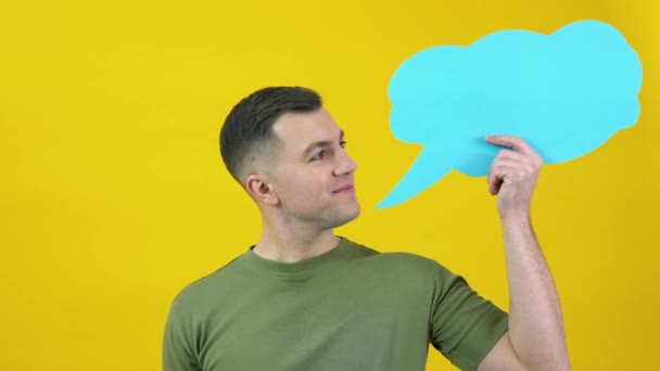 Caucasian middle-aged man in a green T-shirt holding a cloud for the insert text and smiling in the frame. The guy is standing in front of a yellow background with a blue poster for text in his hand. — Vídeo de stock