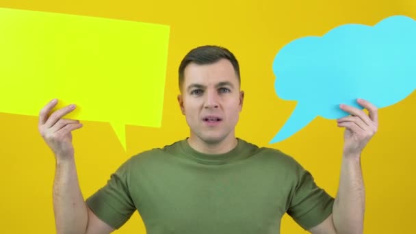 A man holds two backgrounds for the insert text and shouts loudly in the frame. The guy is standing in front of a yellow background with yellow and blue posters for text in his hands — Vídeos de Stock