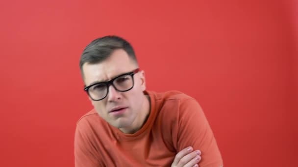 A man in glasses looks into the camera as if he cant see well. The guy in red clothes is standing in front of a red background — Vídeo de Stock