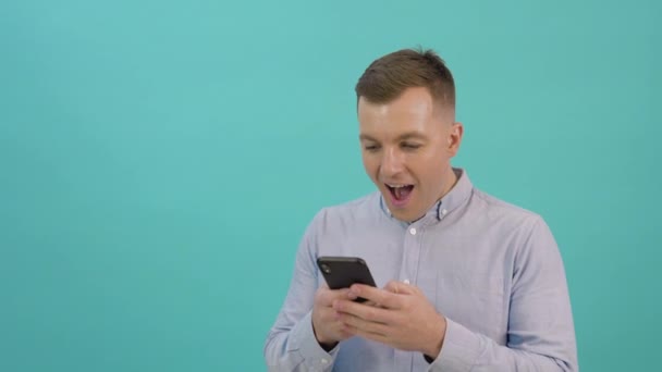 A man in a blue shirt is corresponding on a smartphone and expresses great joy and surprise. Office worker with a mobile phone in his hands stands in front of a bright blue background – Stock-video
