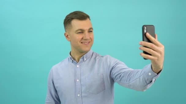 Caucásico hombre de mediana edad en una camisa azul es la videollamada en su teléfono inteligente y expresar alegría. Trabajador de oficina con un teléfono móvil en la mano se para frente a un fondo azul brillante — Vídeos de Stock