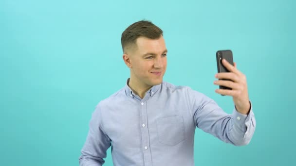 Portrait of positive man in a blue shirt takes a selfie with the emotion of happiness and victory. Office worker with a mobile phone in his hand stands in front of a bright blue background – Stock-video