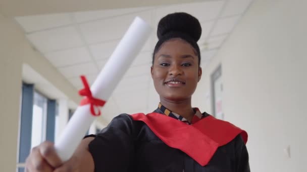 맨틀에서 졸업 한 아프리카 계 미국인 여성은 손에 졸업장을 쥐고 미소를 지으며 서 있습니다. IT 대학교 졸업장을 받고 졸업하다 — 비디오