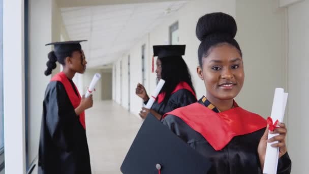 En glædelig kvindelig kandidat med et eksamensbevis i hånden står på universitetet på baggrund af kandidater taler med hinanden. Studenterudvekslingsprogram mellem universiteter rundt om i verden – Stock-video