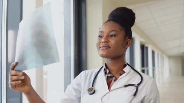 Kadın Afro-Amerikan doktor ya da stajyer akciğerlerinin röntgenini çeker, kapalı alanda tutarak. Uzman, göğsünün şeffaf görüntüsünü kollarında tutuyor ve dikkatlice araştırıyor. — Stok video