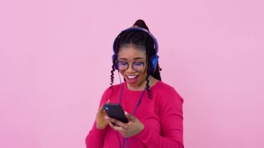 Afrikalı Amerikalı kız fotoğraf stüdyosunda akıllı telefondan müzik dinliyor. Pembe elbiseli genç bir kız, pembe bir geçmişi var.