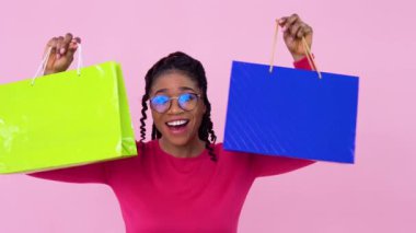 Pembe elbiseli Afro-Amerikalı kız, kağıt kaplamalı çantalarla eğleniyor ve dans ediyor. Pembe arka planda duran genç bir kız.