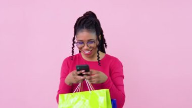 Genç bir kız sağlam bir pembe arka planda duruyor ve online alışveriş yapıyor. Pembe elbiseli neşeli Afro-Amerikalı kadın elinde telefon kullanarak kağıt kaplamalı çantalarla ayakta duruyor.