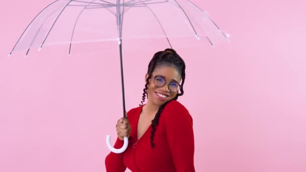 Мила американка-афроамериканка, яка розважається прозорою парасолькою на фотостудії. Підліток у червоному одязі тримає парасольку на твердому рожевому фоні. — стокове відео