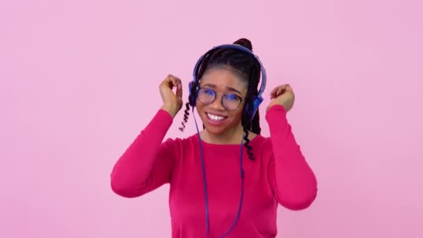 可爱的年轻非洲裔美国音乐爱好者女孩正在照片演播室里听智能手机里的音乐。穿着粉色衣服的少女在坚实的粉色背景上跳舞 — 图库视频影像