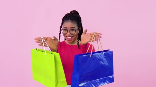 Jovem menina afro-americana alegre em roupas rosa se divertindo e dançando com sacos de papel laminado com alças. Menina adolescente em pé sobre um fundo rosa sólido — Vídeo de Stock