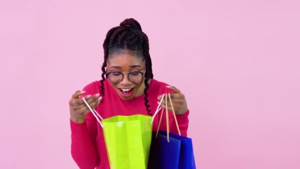 Linda chica afroamericana alegre joven en ropas de color rosa se ve en bolsas laminadas de papel con asas. Adolescente chica de pie sobre un sólido fondo rosa — Vídeo de stock