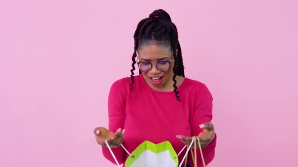 Jeune fille afro-américaine en vêtements roses regarde dans des sacs stratifiés en papier avec des poignées. adolescent fille debout sur un solide rose fond — Video