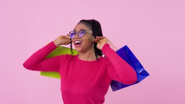 Linda chica afroamericana alegre joven en ropa rosa divertirse y bailar con bolsas laminadas de papel con asas. Adolescente chica de pie sobre un sólido fondo rosa — Vídeos de Stock