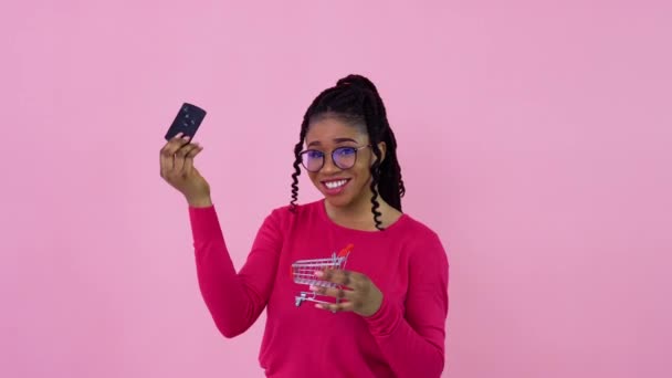 ピンクの服を着たかわいい陽気な若いアフリカ系アメリカ人の女の子が車の鍵を握っておもちゃのショッピングバスケットに入れます。ティーン女の子主婦初心者上のA固体ピンクの背景 — ストック動画