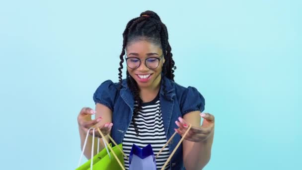 Mignon joyeux jeune fille afro-américaine en vêtements bleus regarde dans des sacs en carton stratifié. adolescent fille debout sur un solide lumière bleu fond — Video