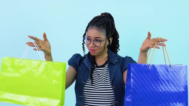 Joyeux jeune fille afro-américaine en vêtements bleus tenant des sacs en papier feuilleté avec des stylos. adolescent fille debout sur un solide lumière bleu fond — Video