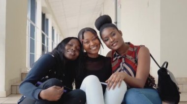 Bir grup Afrikalı Amerikalı öğrenci üniversitede kitaplarla ve tabletle yerde oturup kameraya bakıyorlar. Eğitim kavramı