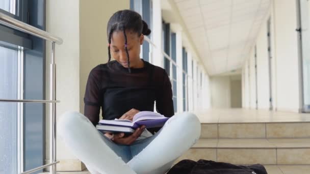 年轻的非洲女学生坐在地板上，手里拿着一本现代图书馆的书 — 图库视频影像