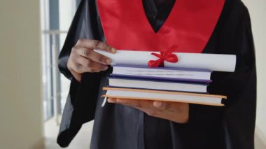 Manto mezunu Afrika kökenli Amerikalı kadın diploması ve elinde kitaplarla duruyor. Yakın çekim.