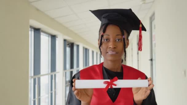 Afrikansk amerikansk kvindelig kandidat i kappe står med et diplom i hænderne og smiler. Kandidateksamen fra et økonomisk universitet med eksamensbevis – Stock-video