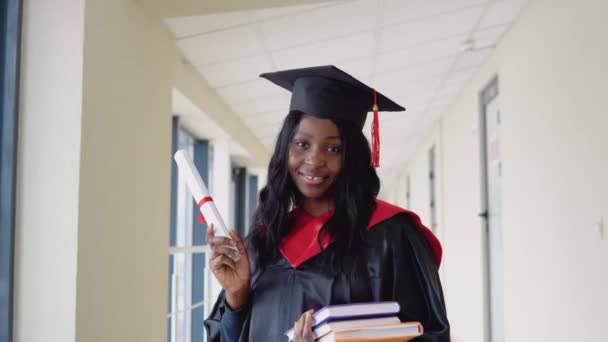 Mujer afroamericana graduada en manto tiene un diploma y libros en sus manos y sonríe. Graduado de una universidad médica con diploma — Vídeo de stock