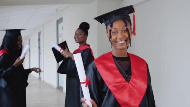 Uma mulher alegre graduada com um diploma na mão está na universidade contra o pano de fundo de graduados conversando uns com os outros — Vídeo de Stock