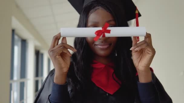 Una mujer graduada con un vestido negro se cubre los ojos con un diploma. Estudiante de posgrado afroamericano en la universidad — Vídeos de Stock