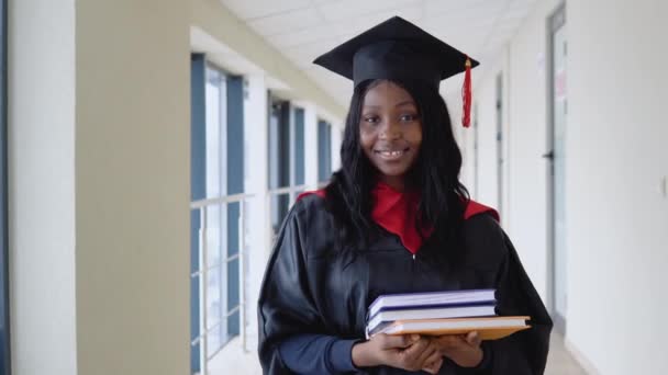 Mujer afroamericana graduada en manto tiene un libro en sus manos y sonríe. Graduado de una universidad médica con diploma — Vídeo de stock