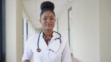 Gülümseyen genç Afrikalı kadın doktor portreyi yakından çek. Mutlu Afro-Amerikan kadın doktor ya da hemşire. Steteskopla poz verip klinikteki kameraya bakıyor.