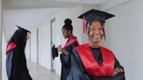Una graduada alegre con un diploma en la mano se levanta en la universidad contra el fondo de graduados que hablan entre sí — Vídeo de stock