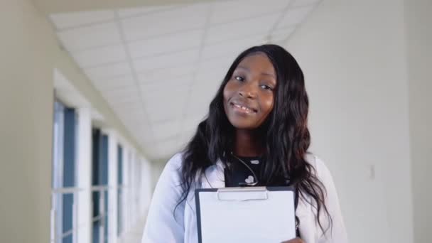 若いアフリカの女性医師の笑顔の肖像画を閉じます。幸せなアフリカ系アメリカ人女性医師または看護師専門家一般開業医ポーズで聴診器を見ながらカメラでクリニック — ストック動画