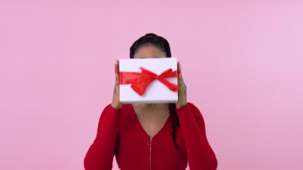 Linda chica afroamericana joven se esconde y mira hacia fuera desde detrás de una caja de regalo. Adolescente chica en ropa roja sostiene una caja blanca con una cinta roja — Vídeo de stock