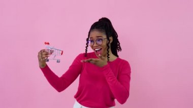Pembe elbiseli neşeli Afro-Amerikalı kız elinde oyuncak bir alışveriş arabası tutuyor. Genç kız ev hanımı acemi, pembe arka planda duruyor.