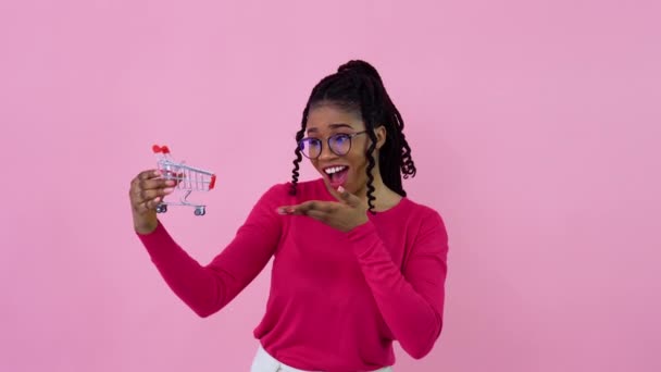 Alegre joven afroamericana chica en ropa rosa sosteniendo un juguete mini carrito de compras. Adolescente chica ama de casa principiante de pie sobre un fondo rosa sólido — Vídeo de stock