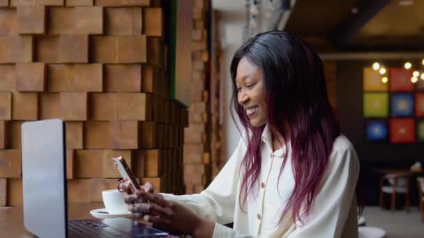 Молодая африканка пользуется телефоном, сидя в кафе с ноутбуком — стоковое видео