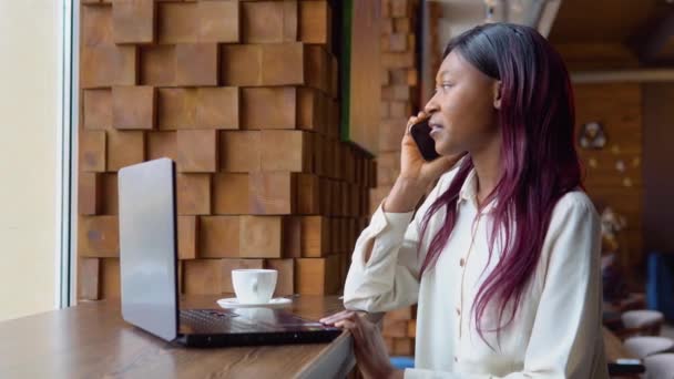 Young black woman talking on the phone while sitting in a cafe with a laptop — Stock Video