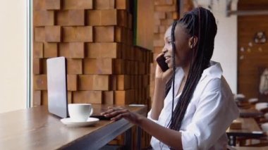 Genç siyahi kadın bir kafede dizüstü bilgisayarla konuşurken telefonla konuşuyor.