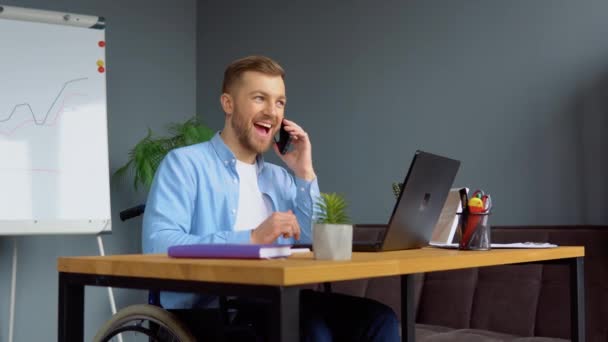 Zmotywowany wesoły niepełnosprawny wolny strzelec rozmawiający przez telefon i robiący notatki siedząc na wózku inwalidzkim w biurze. Koncepcja pracy odległej i niepełnosprawności — Wideo stockowe