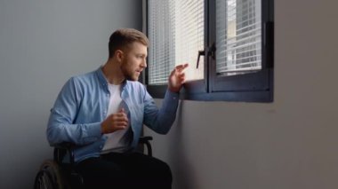 Pencerenin yanında tekerlekli sandalyede oturan üzgün engelli adam.