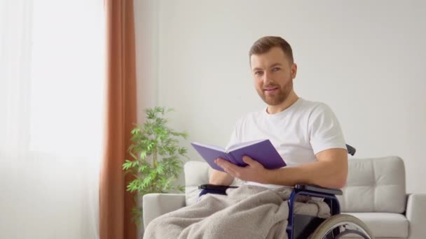 Fröhlich fröhlicher Behinderter liest im Rollstuhl ein Buch und blickt in die Kamera — Stockvideo