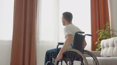 Büyük pencerenin önünde tekerlekli sandalyede oturan beyaz bir adamın arka manzarası. Yalnız genç bir adam evde tek başına bir gün geçiriyor. Engelli konsepti