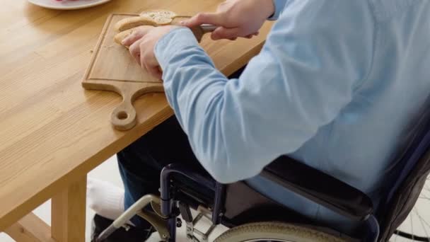Κοντινό πλάνο ενός ανάπηρου σε αναπηρική καρέκλα που κόβει ψωμί στην κουζίνα — Αρχείο Βίντεο